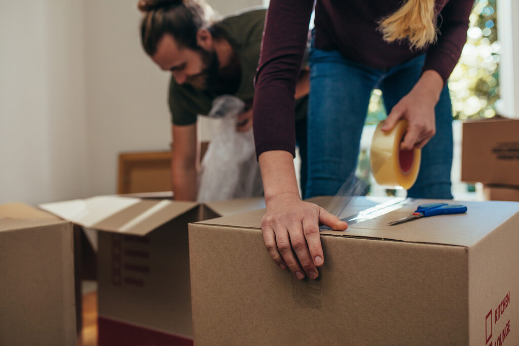 Woman taping a moving box with another person packing a box in the background.