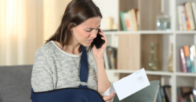 Distressed woman with arm in a sling holding a piece of paper while talking on phone.