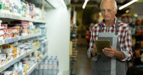 Senior male supermarket owner using a digital tablet while standing in the dairy aisle of his grocery store.