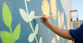 Image of an arm holding a brush painting an outdoor community mural.