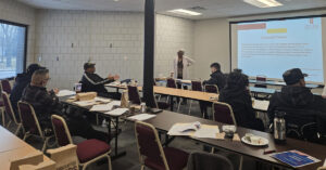 A group of people are sitting in a room with a large screen in front of them.